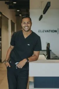 Dr. Brice Gilliam, cosmetic orthodontist at Elevation Orthodontics, standing in modern office setting with logo visible, dressed in professional attire.